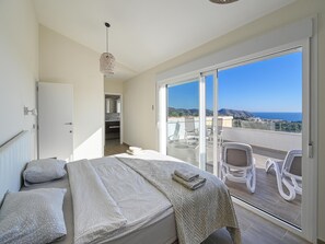 A bedroom with a bed, pillows, a bedside table, and a view of the balcony with chairs.