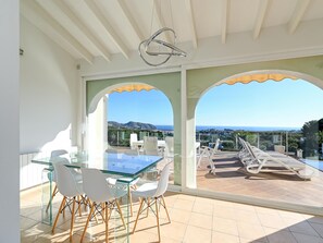 A sunlit terrace with a glass table and chairs, a pool with lounge chairs, and a scenic view of the sea and hills.