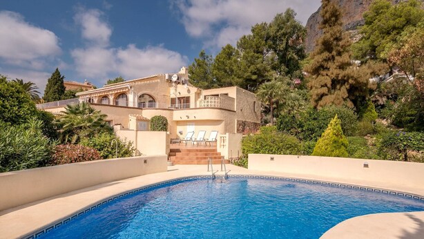 A swimming pool with a clear blue water, surrounded by a beige pool deck. In the background, there is a large building with multiple balconies and a staircase leading to the entrance. - Villa near Montgó with Pool and Beach (Javea)