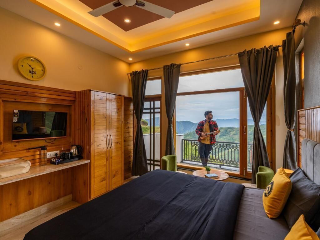 Un dormitorio con cama, televisor, armario de madera, balcón con vistas a las montañas y una persona parada en el balcón.