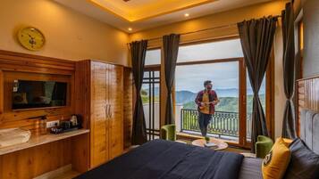 Un dormitorio con cama, televisor, armario de madera, balcón con vistas a las montañas y una persona parada en el balcón.