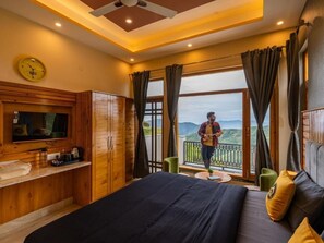 A bedroom with a bed, a TV, a wooden cabinet, a balcony with a view of the mountains, and a person standing on the balcony. - The Hosteller Chakrata (Chakrata)