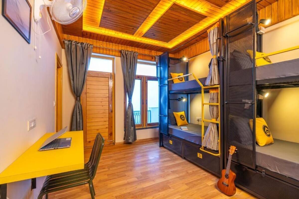 Quarto compacto com beliches, uma pequena mesa com cadeira, um ventilador e um violão.
