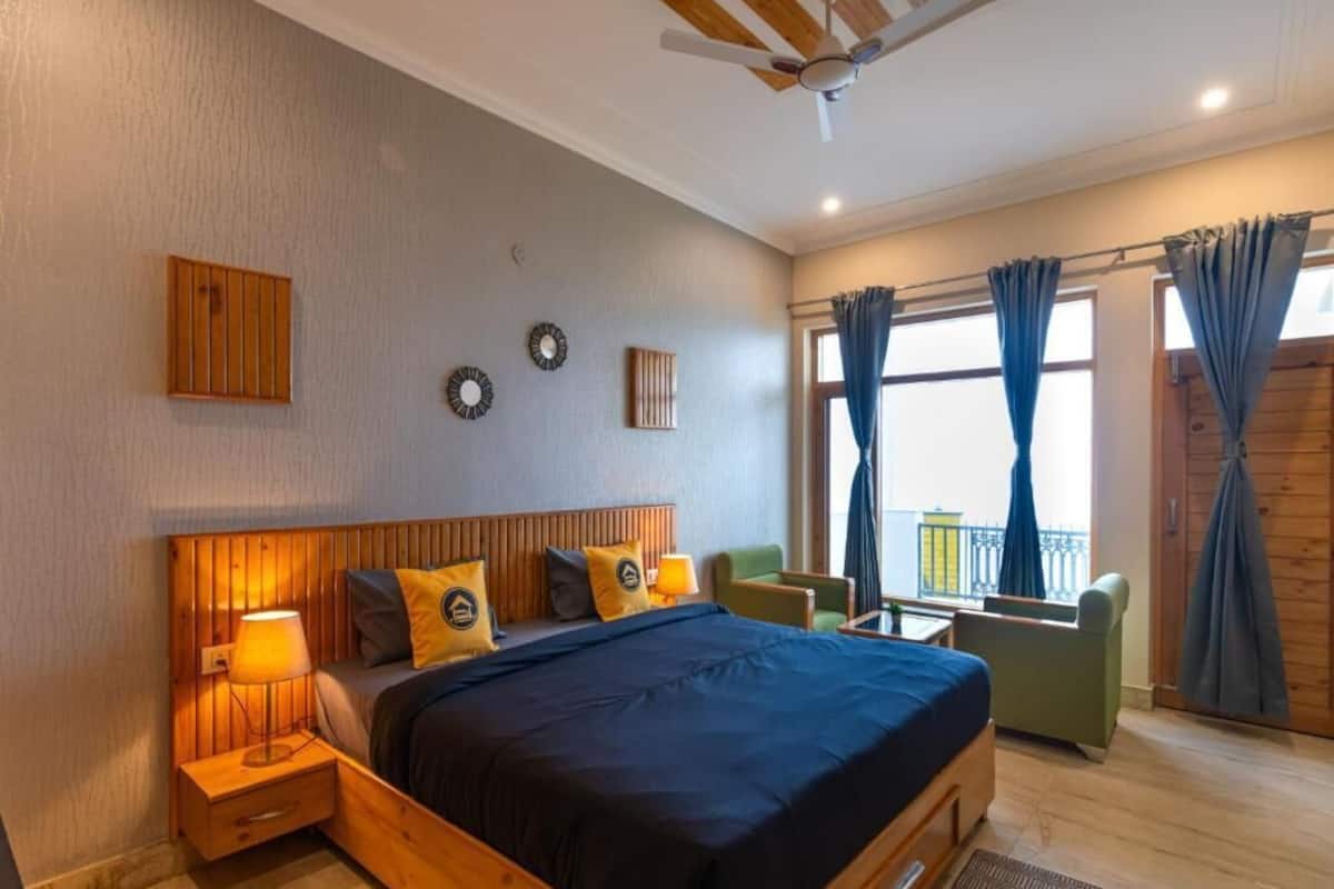 Quarto com cabeceira de madeira, cama com colcha azul escura, duas luminárias de cabeceira, uma pequena mesa com abajur, uma poltrona verde e uma varanda com cortinas.