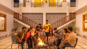 A group of people sitting around a fire pit in a lobby with a staircase and potted plants.