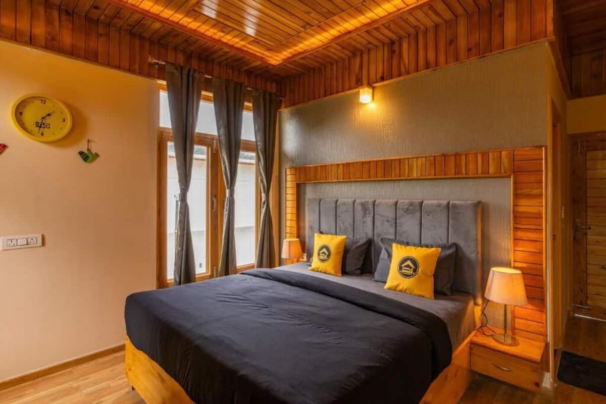 Quarto com teto de madeira, cama com colcha preta e duas almofadas amarelas, mesa de cabeceira com abajur e relógio de parede.