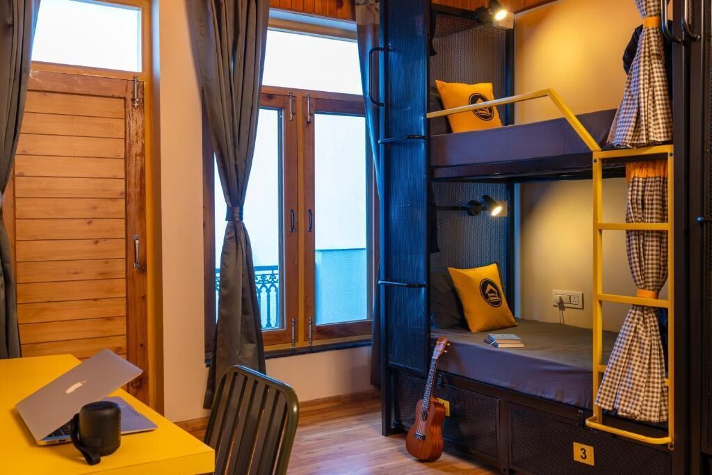 Une petite pièce avec des lits superposés, un bureau avec un ordinateur portable, une guitare et une fenêtre avec des rideaux.