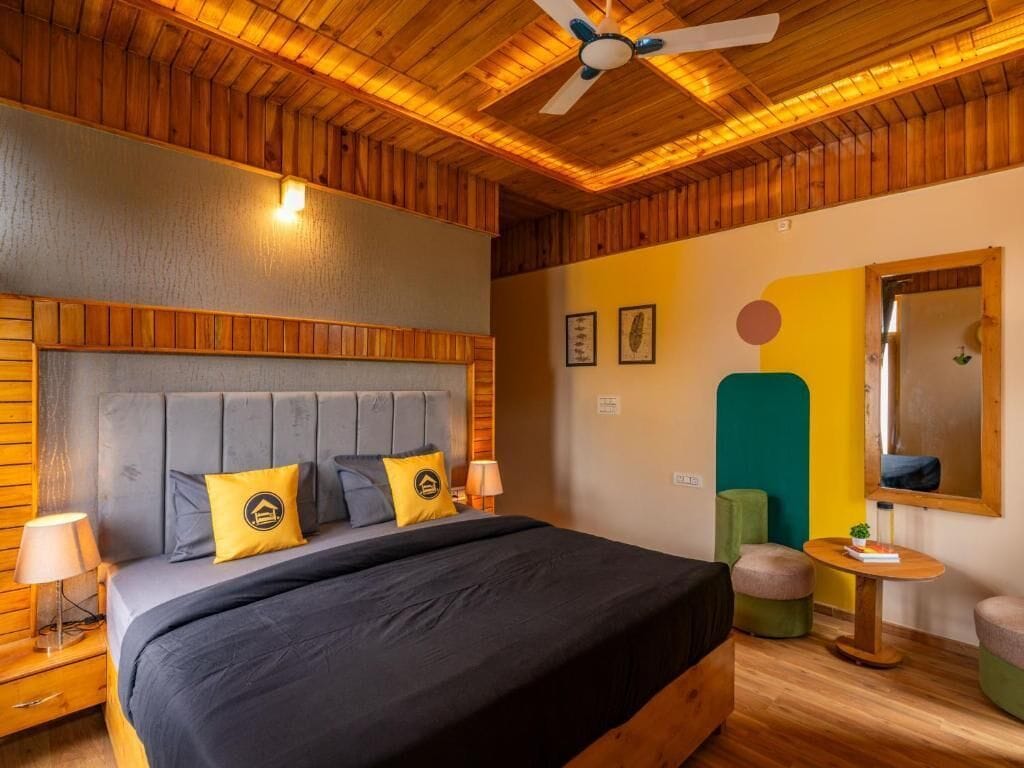 A bedroom with a wooden ceiling, a bed with a grey headboard, and a small table with a lamp.