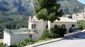 Eine weiße Villa mit Ziegeldach, umgeben von viel Grün und mit Blick auf einen Berg.