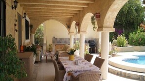 A covered outdoor dining area with wicker furniture, a pool, and potted plants.