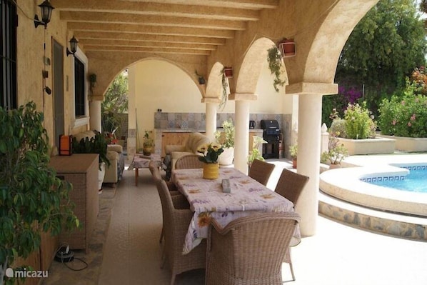 A covered outdoor dining area with wicker furniture, a pool, and potted plants.