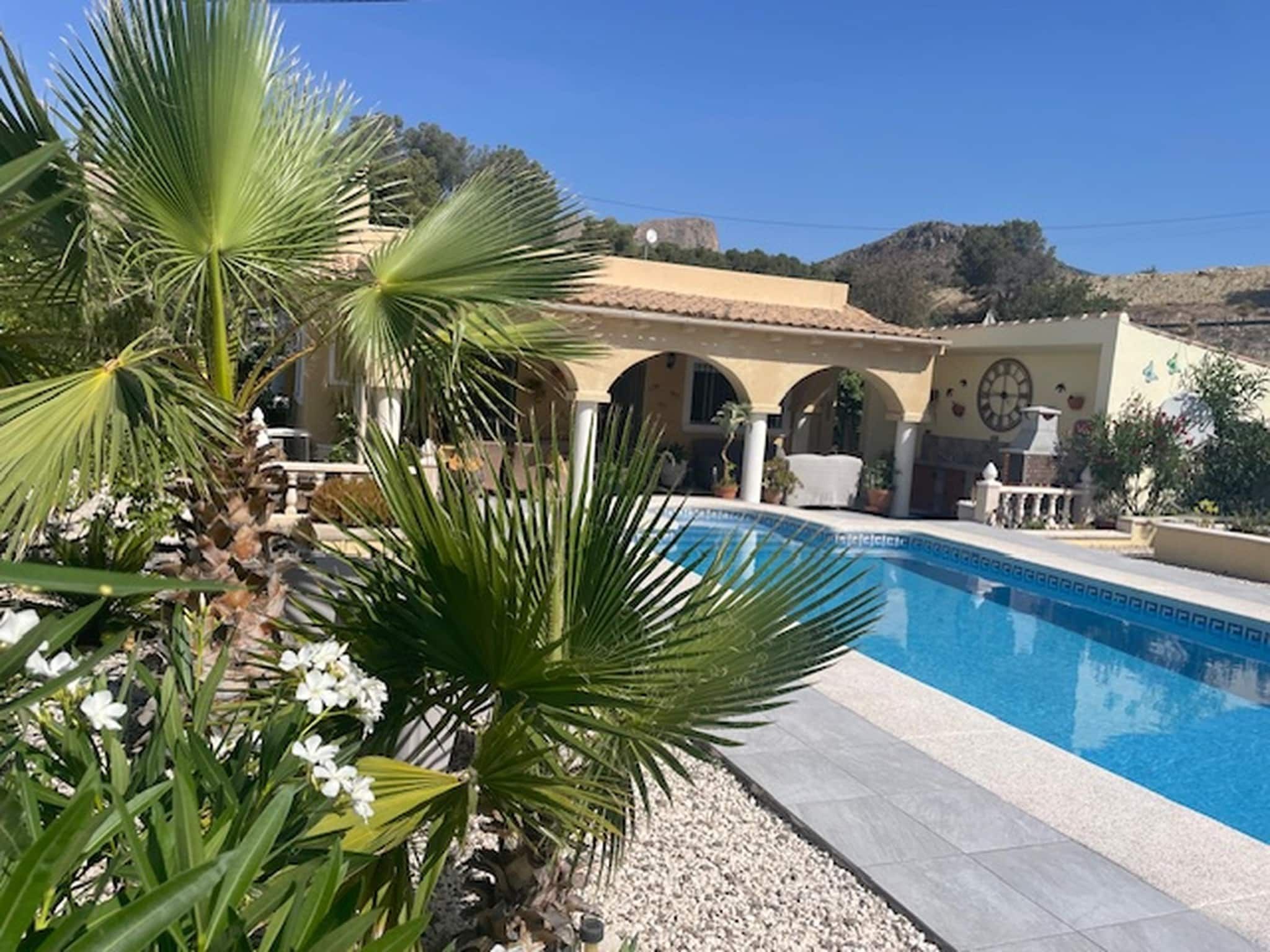 A pool area with palm trees, a building with arches, and a mountainous backdrop.
