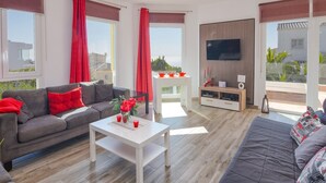 Una sala moderna con un sofá gris, una mesita de centro blanca y un sofá gris. Hay un televisor montado en la pared y una ventana con cortinas rojas.