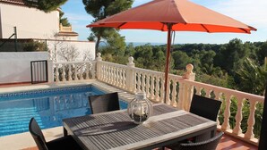 A poolside area with a table and chairs, a red umbrella, and a view of the surrounding landscape.
