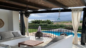 Un área de estar al aire libre techada con vista a la piscina, cojines blancos, una mesa de madera y faroles colgantes.