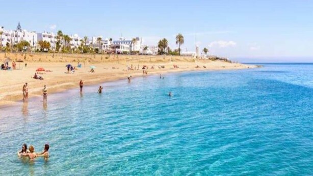 Een strand met helderblauw water, mensen die zwemmen en zonnebaden, en witte gebouwen op de achtergrond.