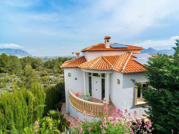 Una casa pequeña y blanca con un balcón curvo, rodeada de abundante vegetación y montañas al fondo.