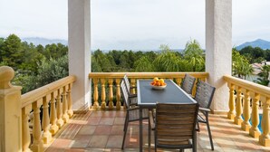 Een balkon met een tafel en stoelen, een fruitschaal en uitzicht op groen en bergen.