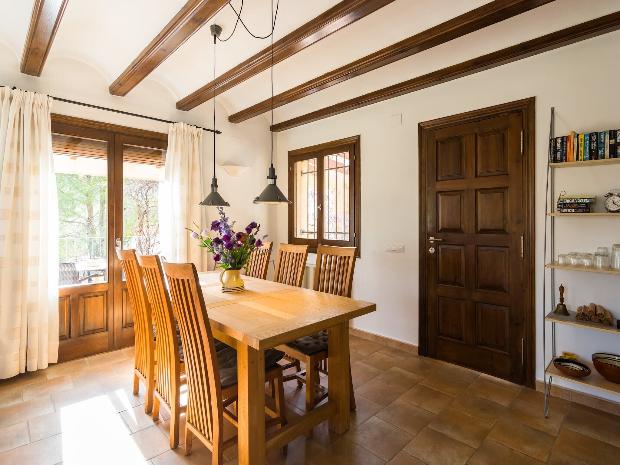 A dining area with a wooden table and chairs, a vase of flowers, and a bookshelf with books and jars.