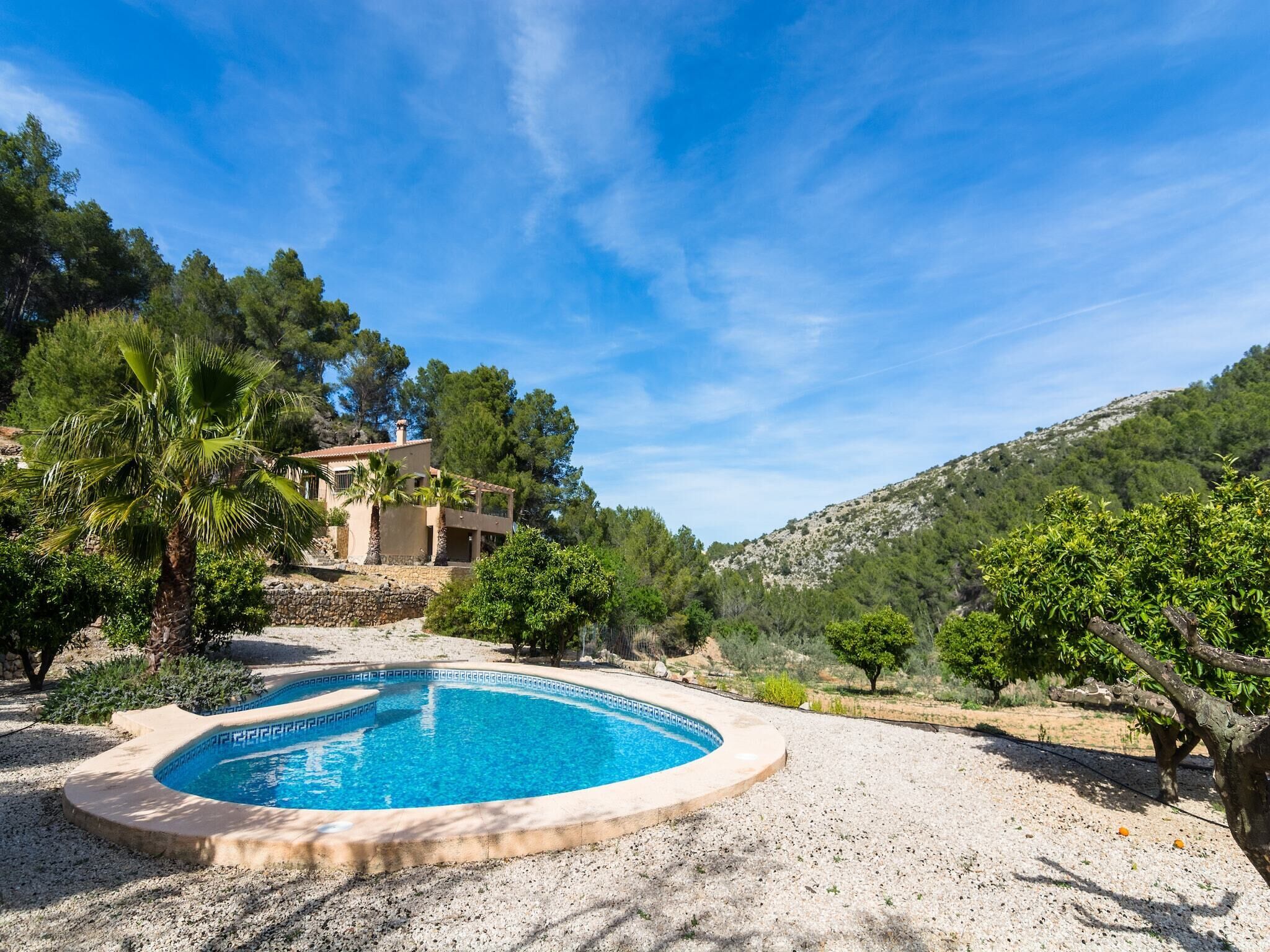 A pool surrounded by a stone patio, with a house in the background and a hilly landscape.