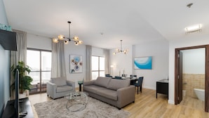 Un salon moderne avec un canapé gris, une table basse et un téléviseur posé sur un meuble noir. On y trouve également un coin repas avec une table et des chaises, ainsi qu’un coin cuisine avec un évier et un plan de travail.