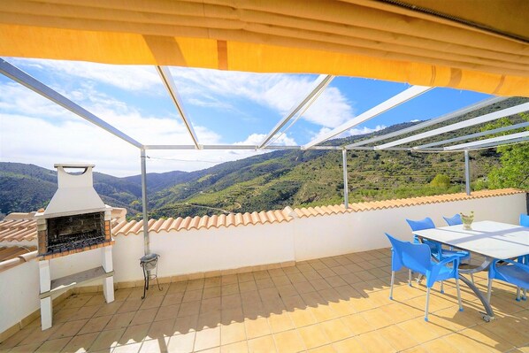 Une terrasse avec un barbecue, des chaises bleues et une table, d’où l’on peut admirer un paysage montagneux.