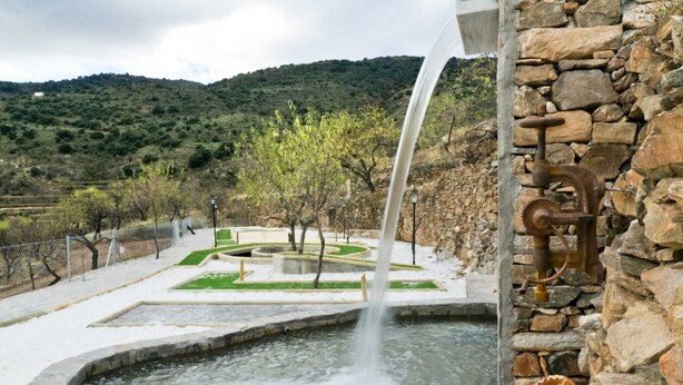 Ein Springbrunnen mit Steinbecken, umgeben von einem angelegten Garten mit Steinmauer und Blick auf eine hügelige Landschaft.
