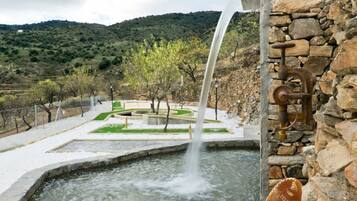 Un élément décoratif composé d’une fontaine en pierre, entouré d’un jardin aménagé avec un mur de pierre et offrant une vue sur un paysage vallonné.