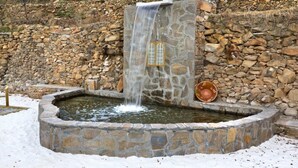 Ein Steinbrunnen mit Wasserfall, umgeben von einer Steinmauer und verschneitem Boden.