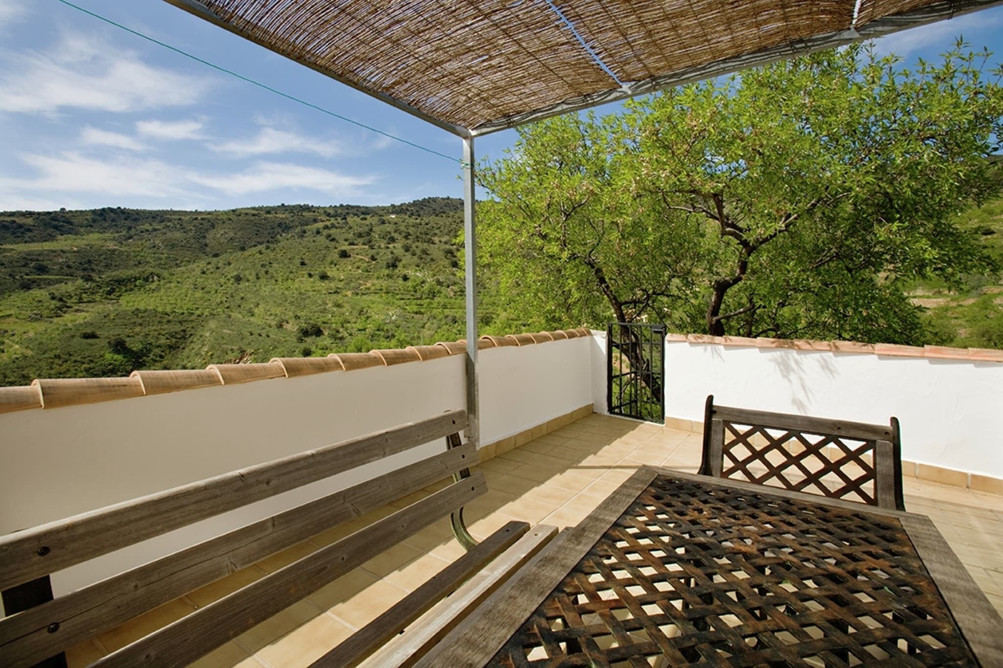 Un balcon doté d’un banc et d’une table en bois, offrant une vue sur un paysage verdoyant, avec des arbres et des collines.