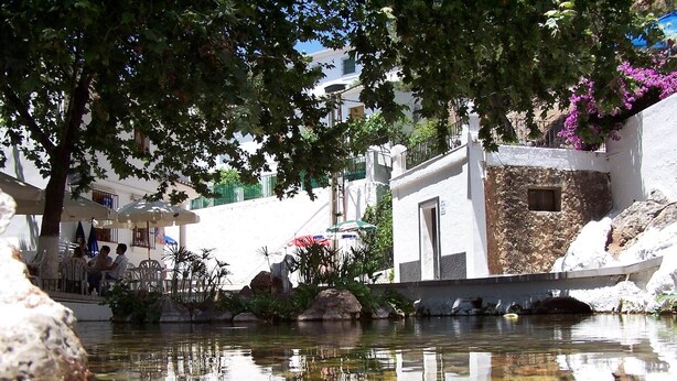 Un entorno exterior tranquilo con una fuente de agua cristalina, muros de piedra y una zona de estar a la sombra de los árboles.