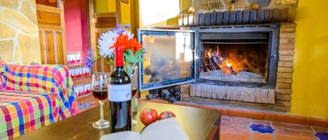 A cozy room with a stone fireplace, a checkered sofa, a table with a wine bottle and book, and a tiled floor.