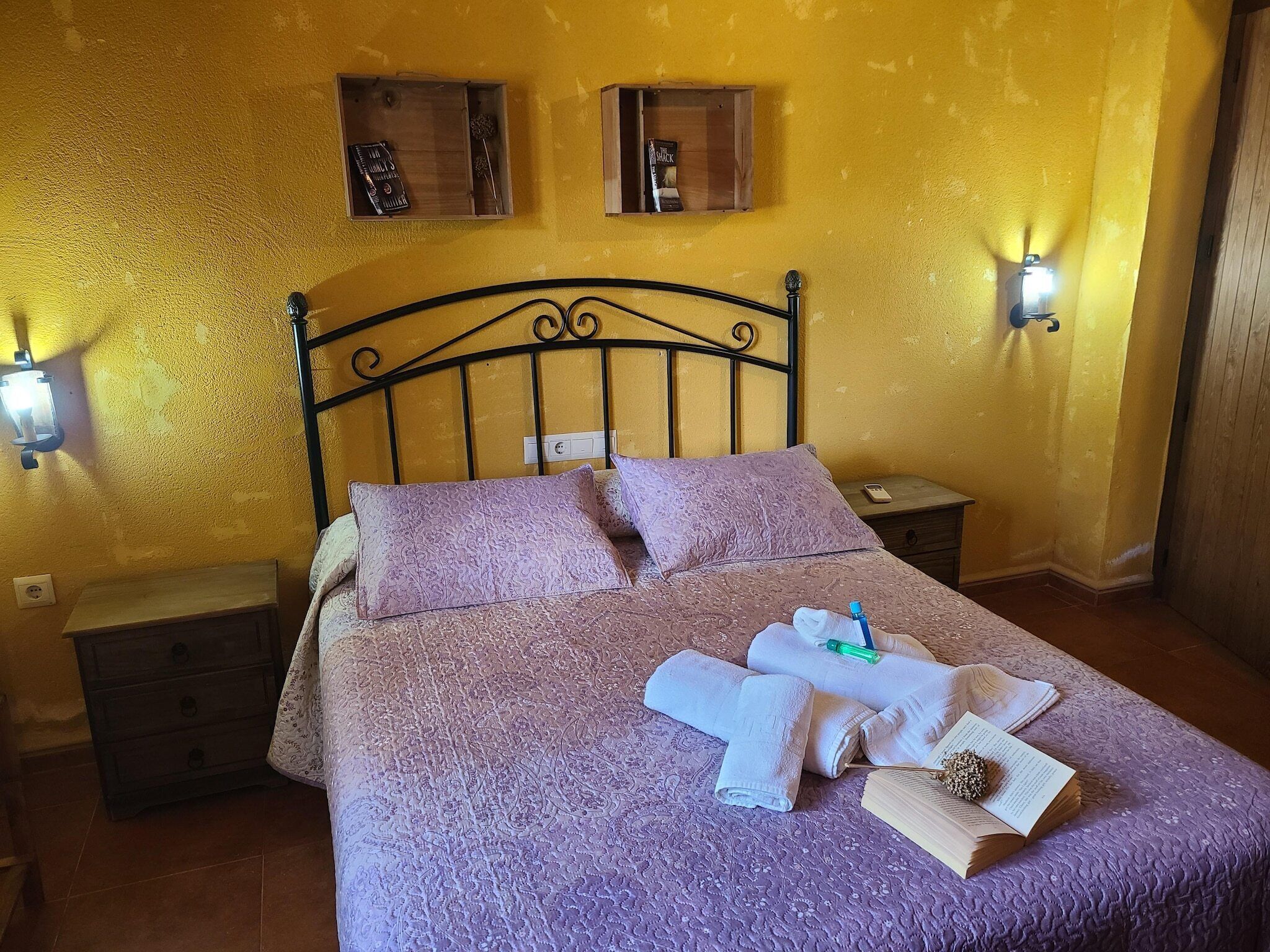 Ein Bett mit gemustertem Überwurf, Kissen und Handtüchern, ein Nachttisch mit Lampe sowie zwei Wandregale mit Büchern und einem dekorativen Gegenstand.