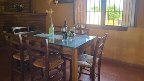 Uma sala de jantar com mesa de vidro, cadeiras de madeira, um vaso com flores secas, uma garrafa de vinho e um copo.