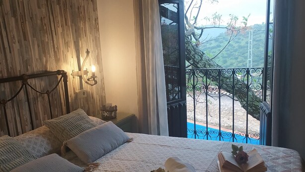 Un dormitorio con cama, almohadas, una mesita de noche, lámpara y vistas a una piscina y colinas.