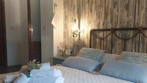 Una cama bien hecha con almohadas y una toalla doblada, una mesita de noche con lámpara, y una lámpara de pared.