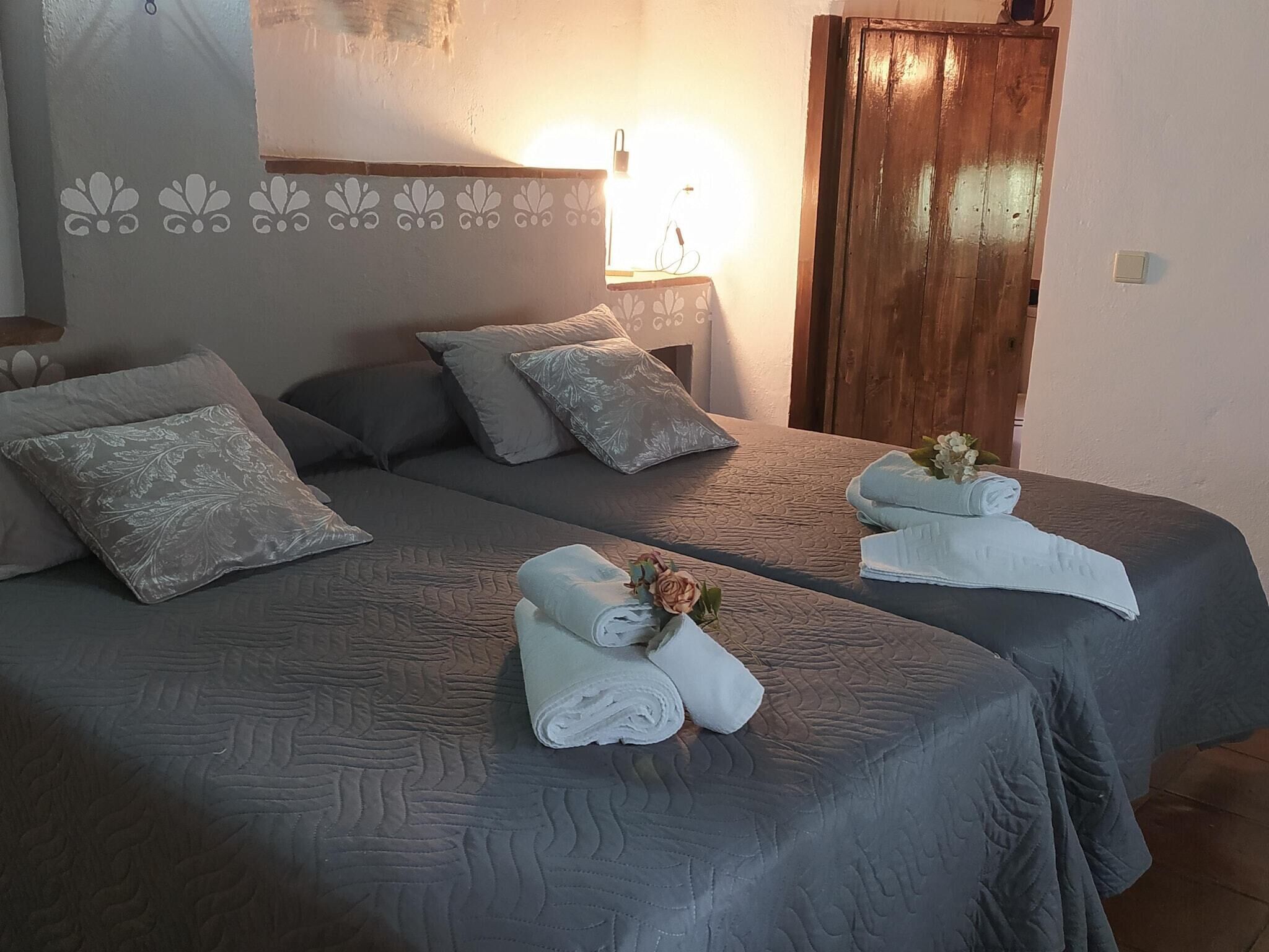 Une chambre d’hôtel avec deux lits, chacun recouvert d’une couverture grise assortie aux oreillers. On y trouve une armoire en bois et un luminaire fixé au mur.