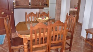 A dining area with a wooden table and chairs, a kitchen in the background, and a brick wall.