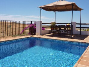 A clear blue pool with a pergola and seating area overlooking a hilly landscape.