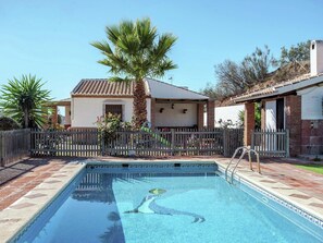 Una piscina con borde de baldosas, cerca de madera y una casa con techo de tejas y una palmera.