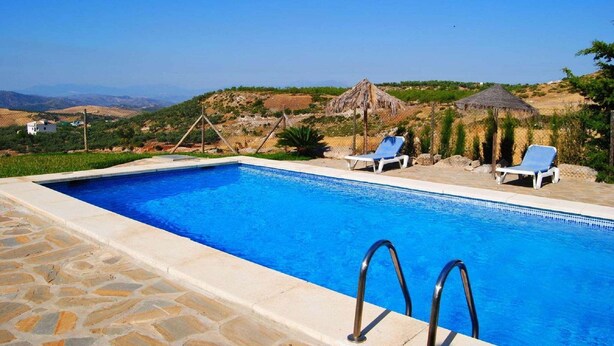 A swimming pool with two lounge chairs and a thatched-roof structure in a hilly landscape.