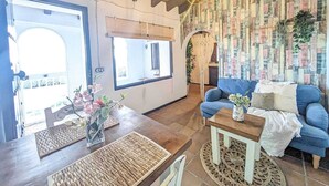 A cozy living room with a blue sofa, wooden coffee table, and a woven rug. There is a large window with a view of the outside, and a patterned wall in the background. - Cottage in La Joya with Pool and Garden (La Joya)