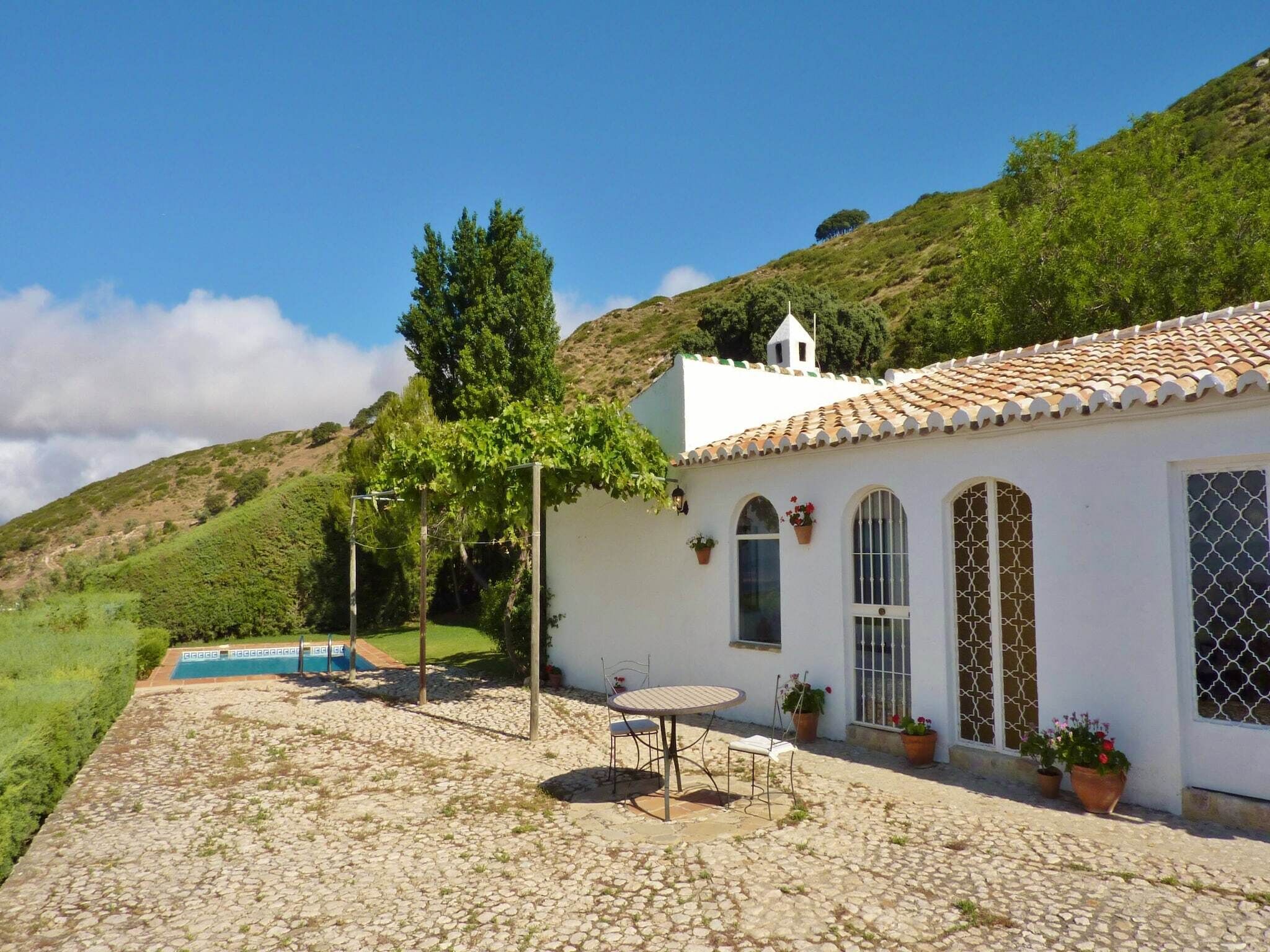 A white building with a tiled roof, a pool, and a patio area with a table and chairs.