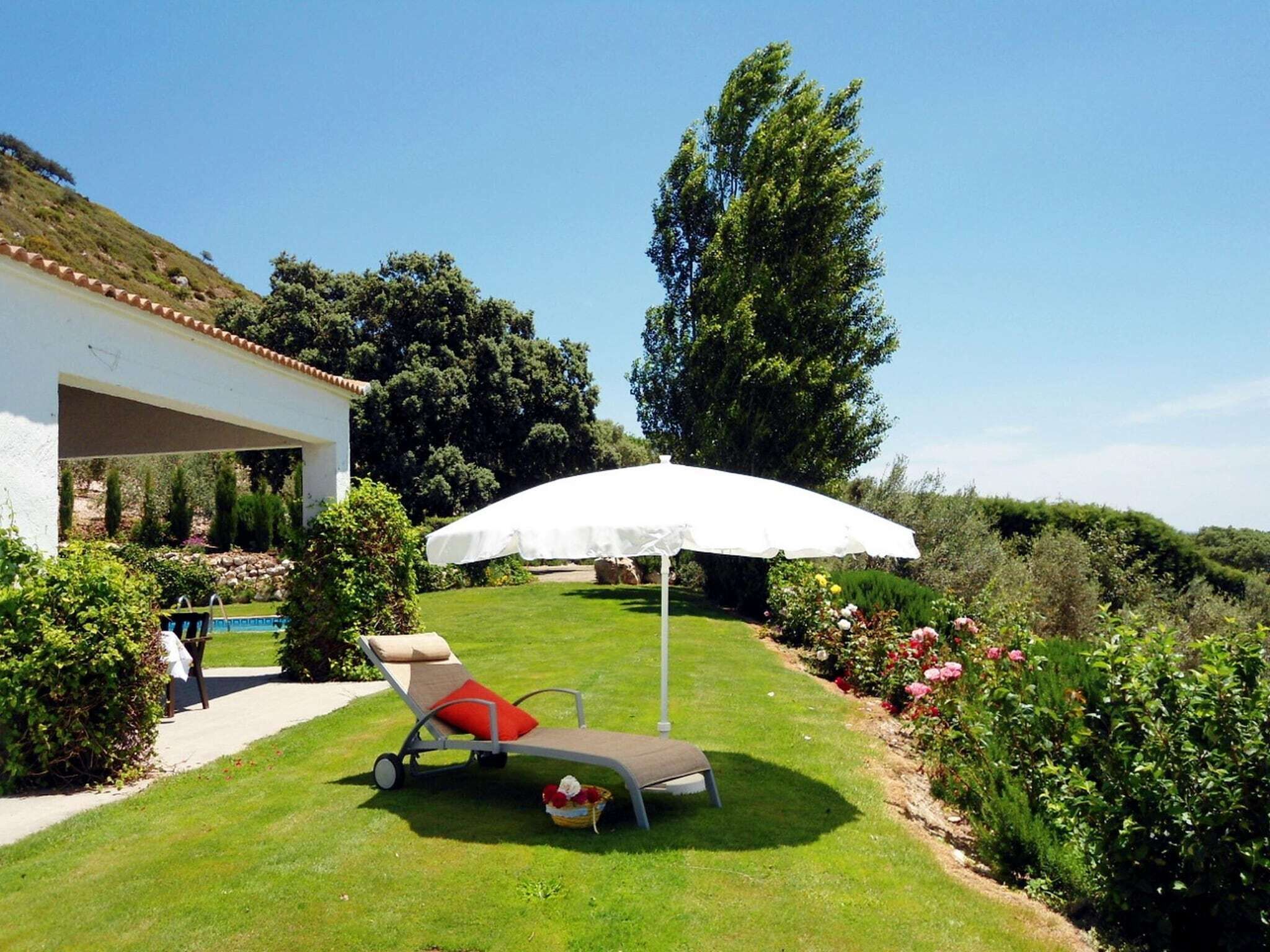 A sun lounger with a white umbrella, a pool, and a garden with various plants and flowers.