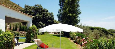 A sun lounger with a white umbrella, a pool, and a garden with various plants and flowers.