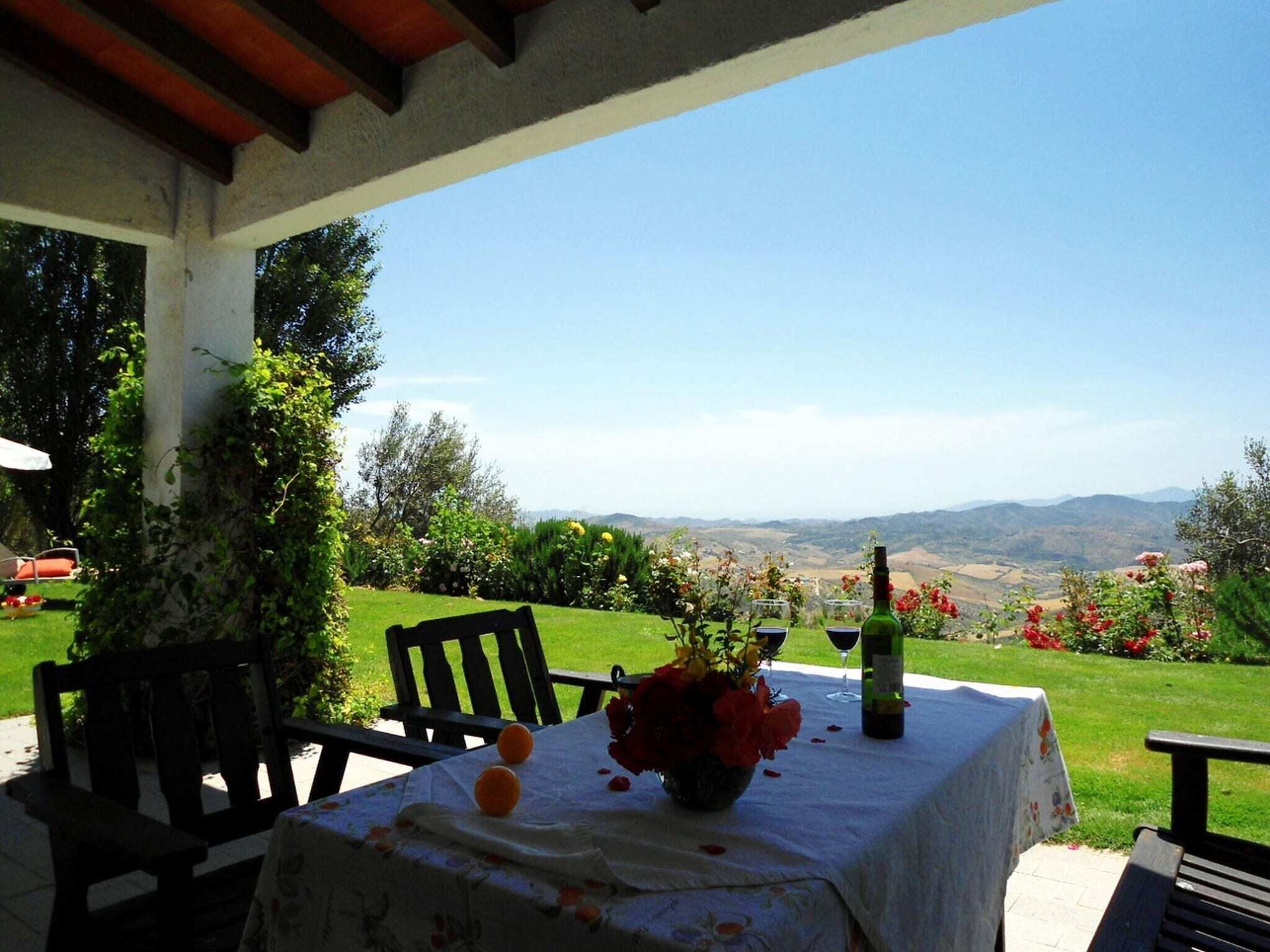 An outdoor patio with a table set for two, featuring a bottle of wine, a vase with flowers, and a tablecloth.