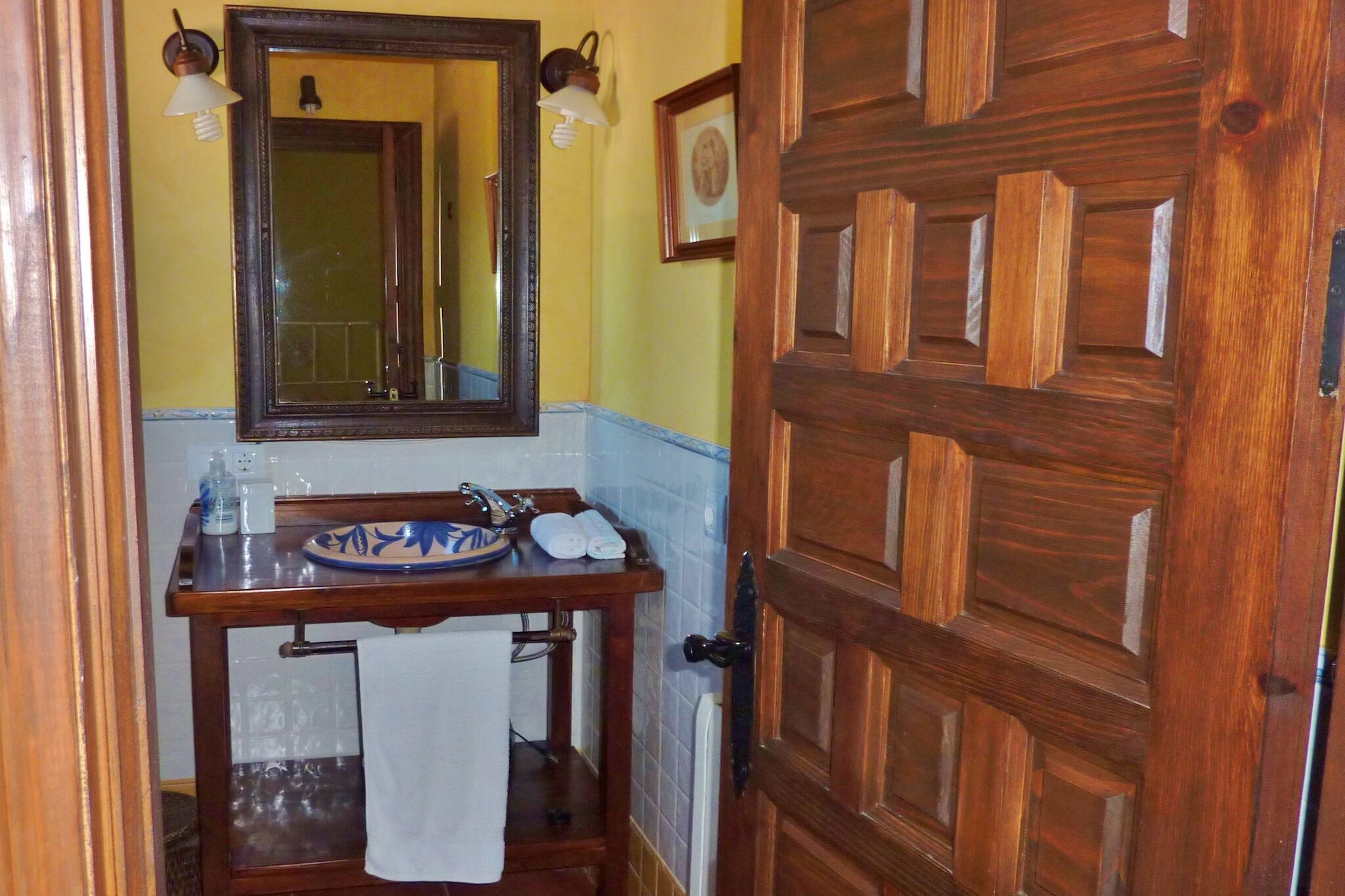 A bathroom with a wooden door, a mirror, a sink with a blue and white ceramic basin, a towel hanging on a rack, and a small shelf with toiletries.