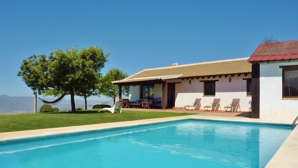 A poolside view of a house with a swimming pool, a hammock, and a patio area with chairs. - Beautiful villa in Andalusia near the forest (La Joya)