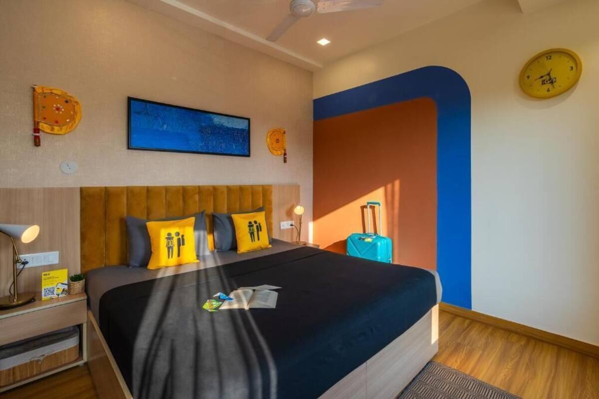A hotel room with a bed, a nightstand, a lamp, a suitcase, and a wall clock.