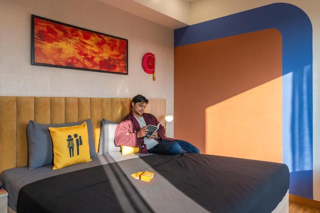 A hotel room with a bed, a reading lamp, a clock, and a painting on the wall.